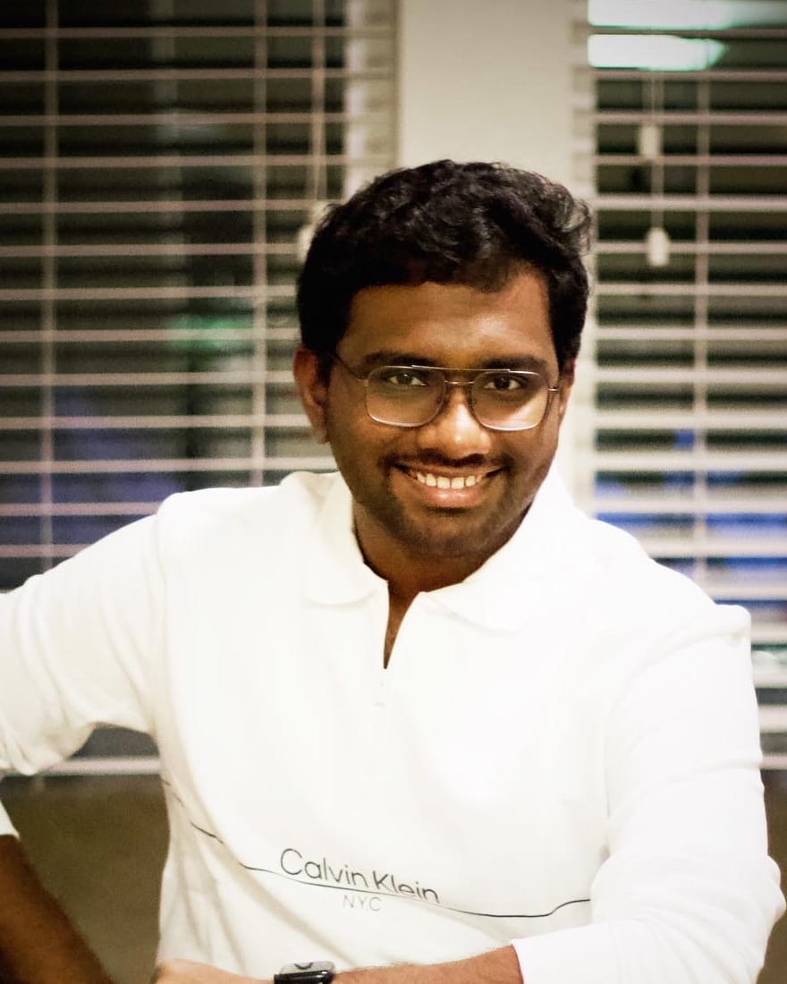 Sai Vivek Peddi, smiling, in a white sweater, sitting in front of window blinds